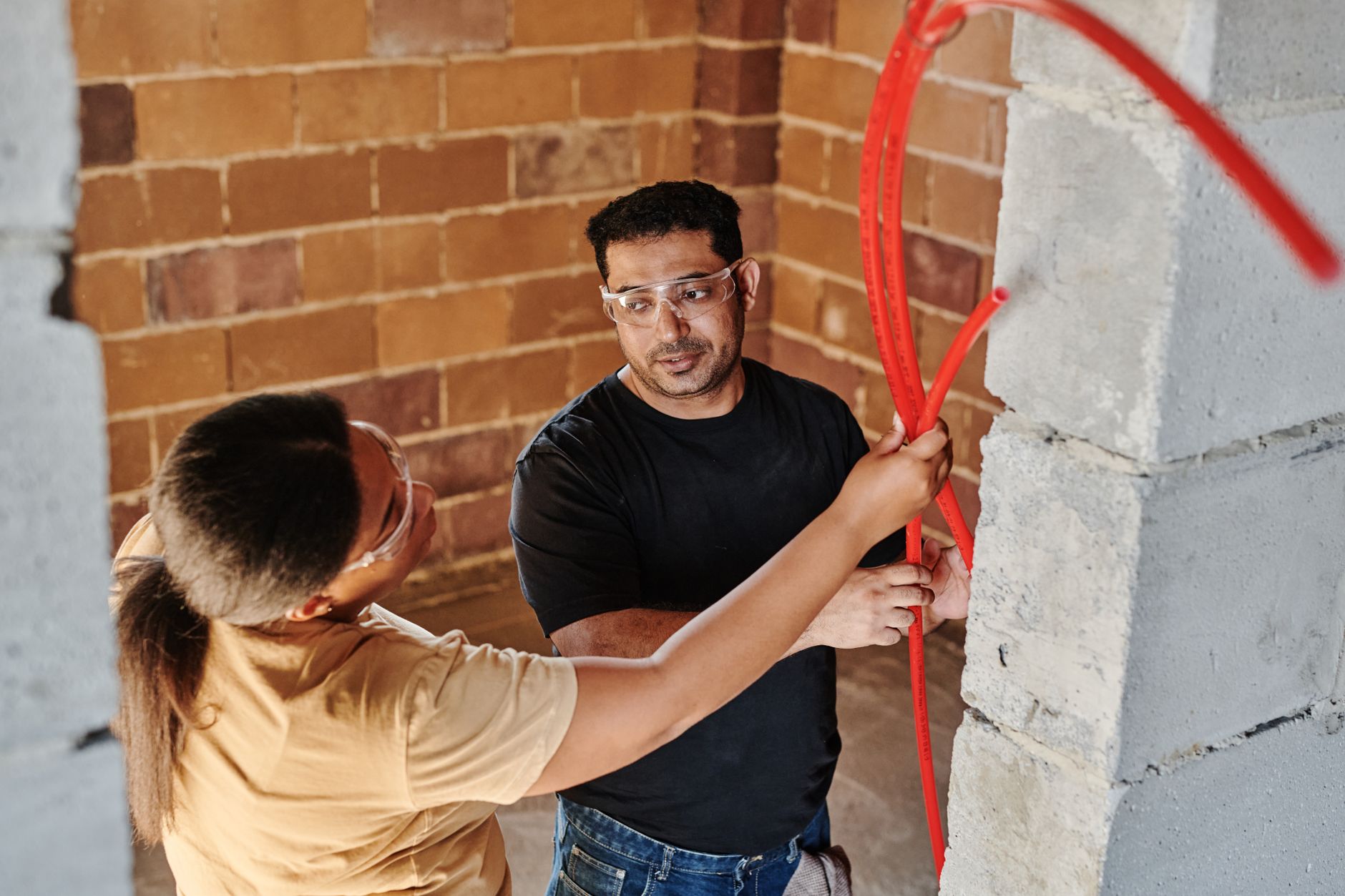 Two workers guiding red tubing through a concrete block wall at a construction site
